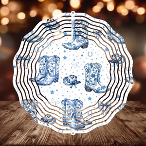 May include: A white metal wind spinner with a blue and white floral pattern. The design features cowboy boots and hats. The spinner has multiple layers and is designed to spin in the wind.