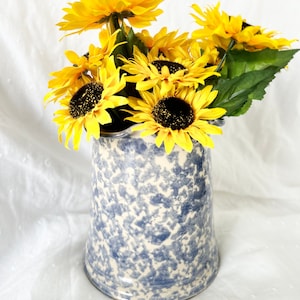 May include: A blue and white speckled ceramic vase filled with bright yellow sunflowers. The vase has a flared top and a speckled pattern. The sunflowers have dark brown centers and green leaves, creating a cheerful floral arrangement.