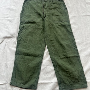 Puede incluir: Pantalones utilitarios de color verde oliva con diseño de cintura alta, cierre de botones y dos grandes bolsillos de parche rectangulares en la parte delantera. Los pantalones tienen un corte recto con costuras visibles en los laterales.