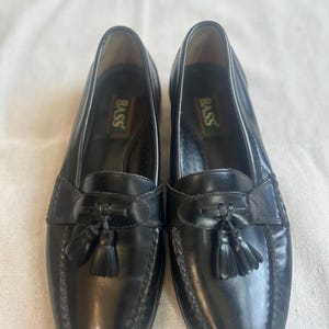 May include: A pair of black leather Bass loafers with a classic design. Each shoe features a decorative tassel and the Bass logo on the insole. The shoes have a glossy finish and a stitched sole.