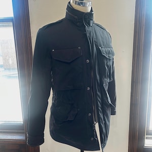 May include: Black jacket with multiple pockets, a high collar, and a zipper closure. The jacket is displayed on a mannequin. The jacket has a military-inspired design with a tailored fit.