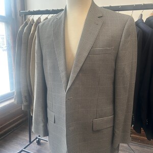 Può includere: Un blazer a quadri grigio e bianco esposto su un manichino. Il blazer ha un risvolto a lancia, una tasca sul petto e due tasche anteriori. Altre giacche sono appese a un attaccapanni sullo sfondo.
