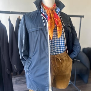 Puede incluir: Un conjunto de moda en un maniquí, que incluye una chaqueta azul marino con cremallera blanca, una camisa a cuadros azul y blanco, pantalones cortos marrones y una bufanda estampada.