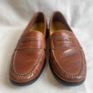 May include: A pair of brown leather penny loafers. The shoes have a classic design with a stitched sole and a leather strap across the vamp. The interior is yellow. The shoes are displayed on a white surface.