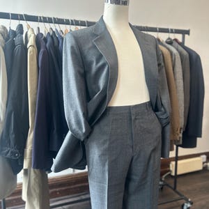 May include: A charcoal gray suit consisting of a single-breasted blazer with notched lapels and matching tailored trousers. The suit is displayed on a mannequin, featuring a smooth fabric texture, structured shoulders, and a clean, classic cut.