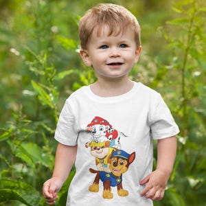 May include: A young child wearing a white t-shirt featuring a colorful graphic of the Paw Patrol characters. The child is smiling and standing in front of a green, leafy background. The shirt design includes images of Chase, Marshall, and other characters.