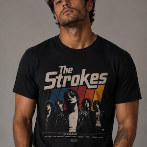 Puede incluir: Camiseta negra con el nombre de la banda "The Strokes" en una fuente retro con un diseño de rayas coloridas. Debajo, una imagen en blanco y negro de los miembros de la banda. También se ve el texto "LIVE IN CONCERT WORLD TOUR".