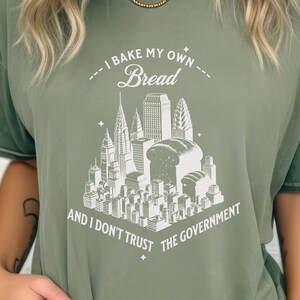May include: A green t-shirt with a white graphic of a city skyline with loaves of bread replacing some of the buildings. The text reads "I bake my own bread and I don't trust the government."