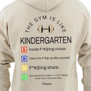 May include: Beige hoodie with the text "THE GYM IS LIKE KINDERGARTEN" and a dumbbell graphic. The back of the hoodie lists gym rules, including "Inside F*@ing voices" and "Clean the F*@ up after yourself."