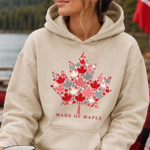 May include: A beige hooded sweatshirt featuring a large graphic of a maple leaf composed of smaller red, white, and grey maple leaf patterns. The text "MADE OF MAPLE" is printed in red below the graphic. The hoodie has a front kangaroo pocket.