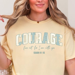 May include: A light yellow t-shirt with the word "COURAGE" in a blue and white block letter design. Below the word "COURAGE" is the text "fear not for I am with you" and "ISAIAH 41:10" in a script font.