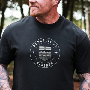 May include: Black t-shirt with a circular graphic reading "REPUBLIC OF ALBERTA" and a stylized emblem. The emblem includes a horizontal stripe, a mountain range, and a shield shape. The shirt is worn by a person.