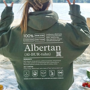 military green Alberta hoodie with black ingredient label style back design reading “Albertan (Al-BUR-tuhn)” featuring prairie values icons, independence themed humor, and Alberta pride text