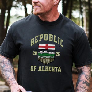 May include: Black t-shirt with the text "REPUBLIC OF ALBERTA" in a vintage style. The shirt features a crest with a red and white flag, mountains, and the numbers "20" and "26". The person is holding a dark blue travel mug.