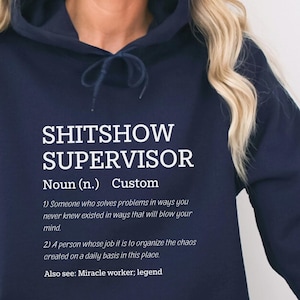 May include: Navy blue hoodie with a white print that reads "SHITSHOW SUPERVISOR" and a definition of the term. The definition includes the text "Custom", "Noun (n.)", "Someone who solves problems in ways you never knew existed in ways that will blow your mind", "A person whose job it is to organize the chaos created on a daily basis in this place", and "Also see: Miracle worker; legend".