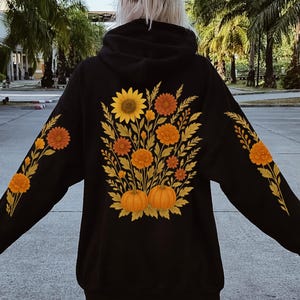 May include: Black hoodie featuring a floral design with sunflowers, orange flowers, and pumpkins. The sleeves have a floral pattern. The design is in shades of yellow, orange, and green.