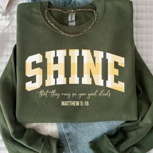 Puede incluir: Sudadera verde oliva con la palabra "SHINE" en letras grandes y blancas con relleno a cuadros amarillos. Debajo está el texto "that they may see your good deeds" y "MATTHEW 5:16". Una cadena dorada está sobre la sudadera.