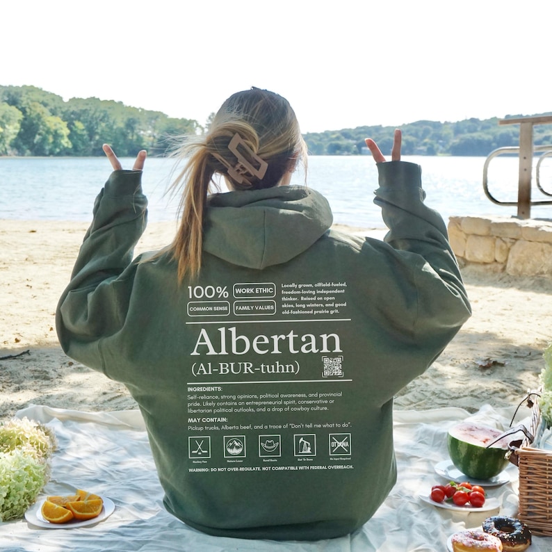 military green Alberta hoodie with black ingredient label style back design reading “Albertan (Al-BUR-tuhn)” featuring prairie values icons, independence themed humor, and Alberta pride text