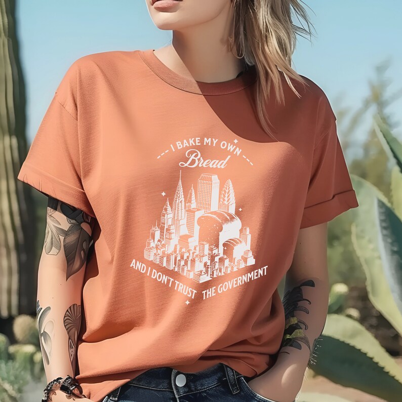 May include: A rust-colored t-shirt with a white graphic of a city skyline with loaves of bread. The text on the shirt reads "I bake my own bread and I don't trust the government."