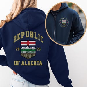 Peut inclure: Sweat à capuche bleu marine avec fermeture éclair, portant l'inscription dorée "REPUBLIC OF ALBERTA". Le dos affiche un écusson avec une croix rouge, des montagnes et les années 20 et 26. Un petit écusson est sur la poitrine.