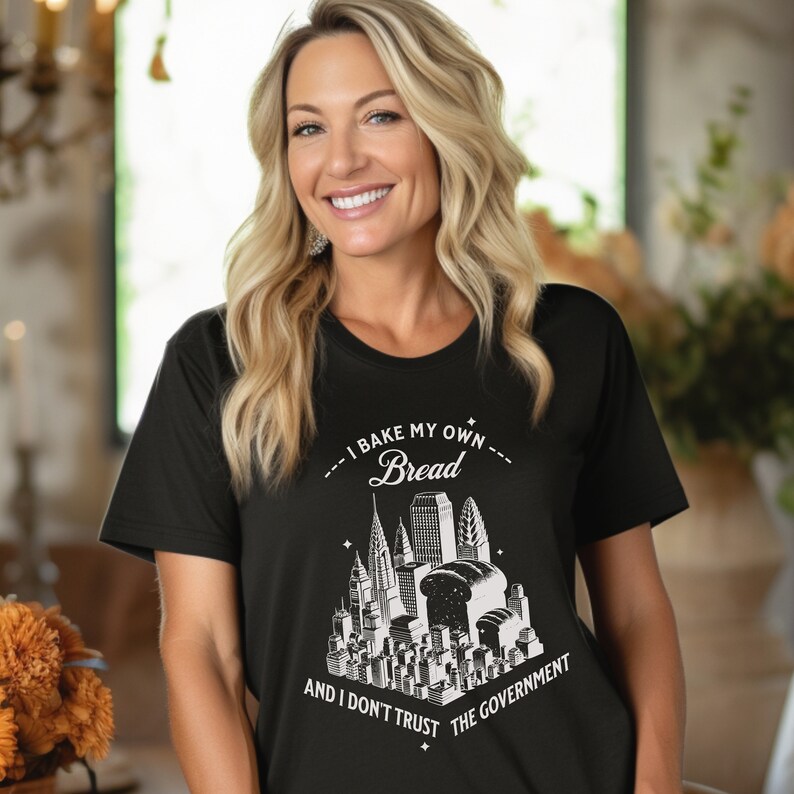 May include: Black t-shirt with a white graphic design of a city skyline with a loaf of bread in the center. The text reads "I bake my own bread and I don't trust the government."