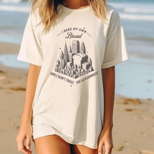 May include: A white t-shirt with a graphic of a city skyline with a loaf of bread in the center. The text on the shirt reads "I bake my own bread and I don't trust the government."