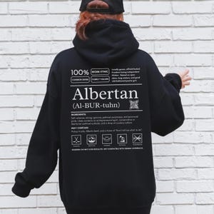 black Alberta hoodie with black ingredient label style back design reading “Albertan (Al-BUR-tuhn)” featuring prairie values icons, independence themed humor, and Alberta pride text