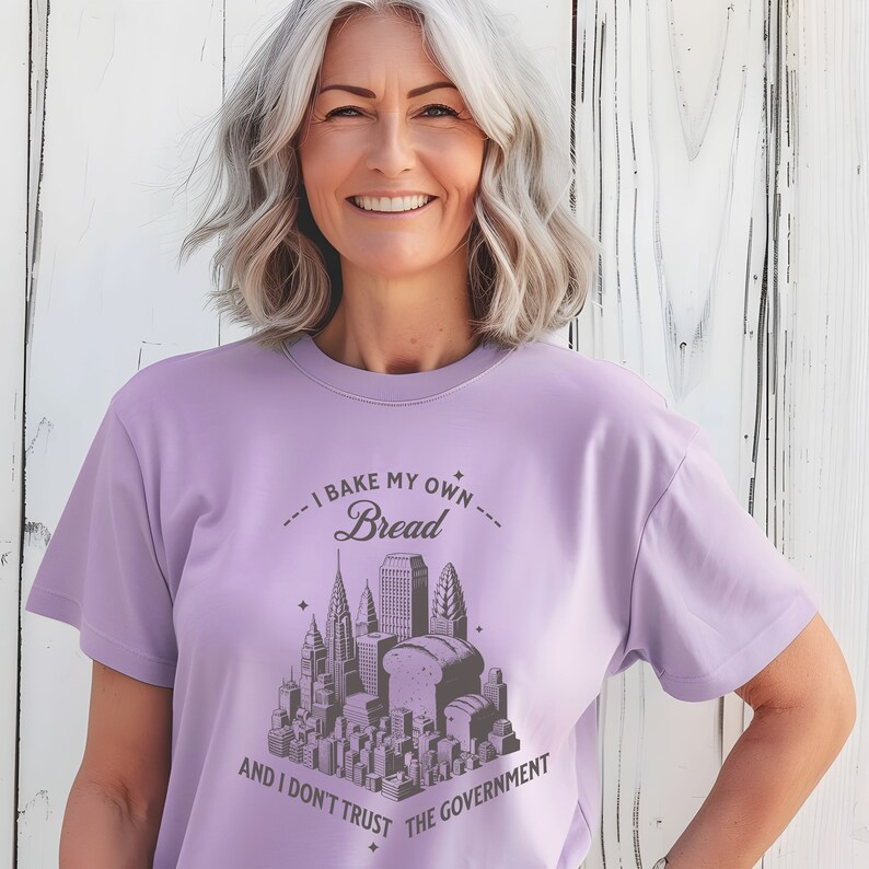 May include: A lavender t-shirt with a graphic of a city skyline with loaves of bread in place of some of the buildings. The text on the shirt reads "I bake my own bread and I don't trust the government."