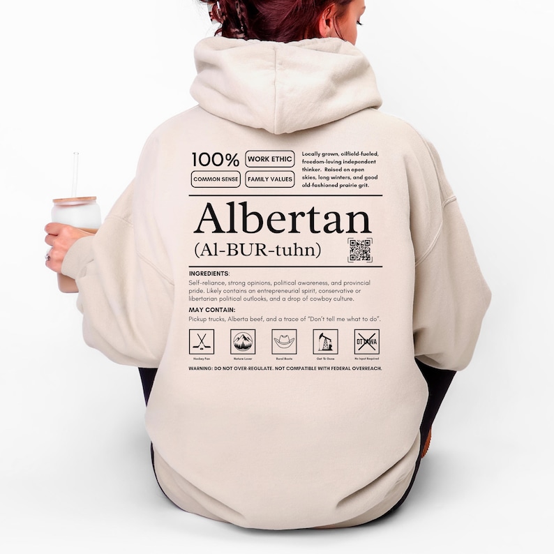 Sand colored (light beige) Alberta hoodie with black ingredient label style back design reading “Albertan (Al-BUR-tuhn)” featuring prairie values icons, independence themed humor, and Alberta pride text