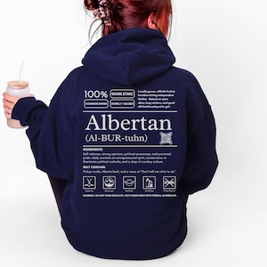 navy blue Alberta hoodie with black ingredient label style back design reading “Albertan (Al-BUR-tuhn)” featuring prairie values icons, independence themed humor, and Alberta pride text
