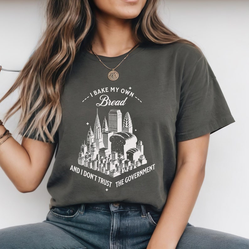 May include: A dark green t-shirt with a white graphic of a city skyline with a loaf of bread in the center. The text on the shirt reads "I bake my own bread and I don't trust the government."