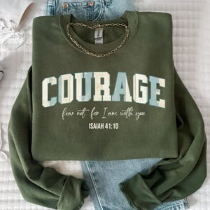 May include: An olive green sweatshirt with the word "COURAGE" in white and light blue lettering. Below the word "COURAGE" is the text "fear not for I am with you" and "ISAIAH 41:10". A gold chain necklace is draped on the sweatshirt.