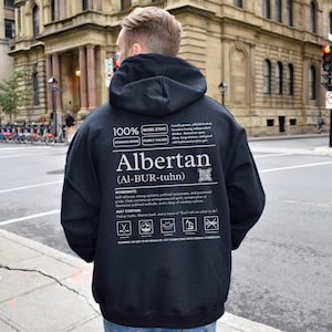 black Alberta hoodie with black ingredient label style back design reading “Albertan (Al-BUR-tuhn)” featuring prairie values icons, independence themed humor, and Alberta pride text