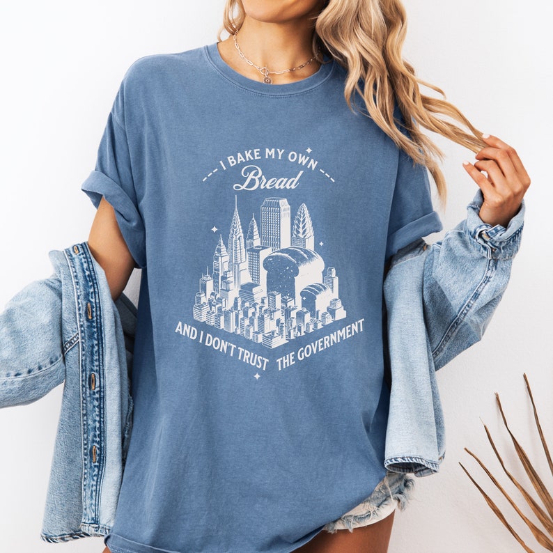 May include: A blue t-shirt with a white graphic of a city skyline with a loaf of bread in the center. The text on the shirt reads "I bake my own bread and I don't trust the government."