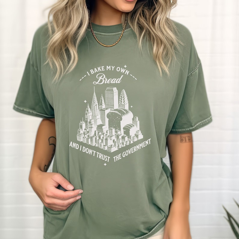 May include: A green t-shirt with a white graphic of a city skyline with loaves of bread replacing some of the buildings. The text reads "I bake my own bread and I don't trust the government."