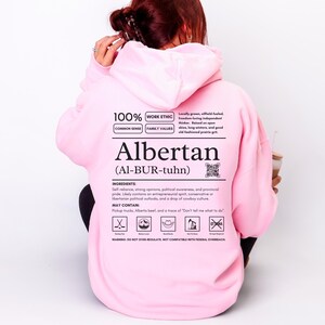 light pink Alberta hoodie with black ingredient label style back design reading “Albertan (Al-BUR-tuhn)” featuring prairie values icons, independence themed humor, and Alberta pride text
