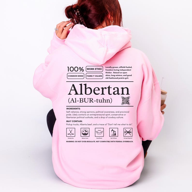 light pink Alberta hoodie with black ingredient label style back design reading “Albertan (Al-BUR-tuhn)” featuring prairie values icons, independence themed humor, and Alberta pride text