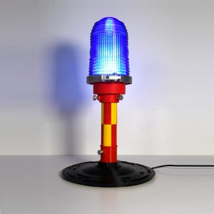 May include: A blue-glowing, cylindrical lamp with a red and yellow striped pole. The lamp has a black base and a silver ring around the blue light. The lamp is plugged into a power outlet. The design resembles an airport runway light.