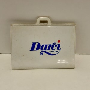May include: A white plastic case with a handle at the top. The case has the blue text "Darci" with a red dot above the "i" and the words "Cover Girl" below the name. The case is rectangular with rounded corners.