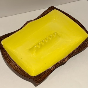 May include: A rectangular ashtray with a bright yellow interior and a textured, brown, wood-grain-like border. The ashtray has a built-in cigarette rest. The design is a mid-century modern style.