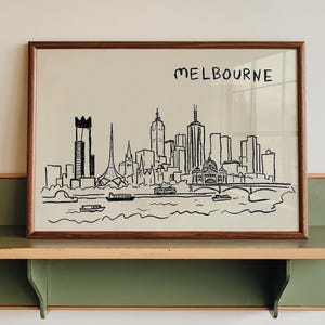 May include: A framed black and white sketch of the Melbourne skyline, with the city name above the image. The artwork is displayed on a green and tan wall shelf.