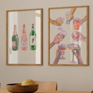 May include: Two framed watercolor prints. One features three wine bottles in pink, green, and green. The other depicts hands holding various cocktail glasses. The prints are displayed above a wooden dining table with a bowl and fruit.