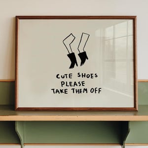 Może przedstawiać: Oprawiony wydruk z brązową drewnianą ramą, przedstawiający czarno-białą ilustrację nóg i butów. Tekst brzmi "CUTE SHOES PLEASE TAKE THEM OFF". Wydruk jest wyświetlany na zielonej i drewnianej półce.