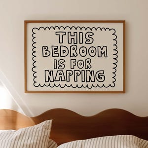 Op de afbeelding: Een slaapkamer met een houten bedframe en gestreepte beddengoed. Een ingelijst bord boven het bed leest "THIS BEDROOM IS FOR NAPPING" in zwarte letters. De kamer heeft een neutraal kleurenpalet.