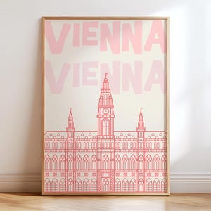 May include: A framed art print featuring a pink line drawing of a building with a clock tower, set against a cream background. The word "VIENNA" is repeated in pink at the top. The artwork is displayed on a wooden floor.