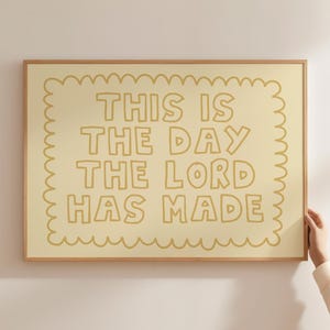 May include: A framed print with the text "THIS IS THE DAY THE LORD HAS MADE" in light brown. The text is on a cream background with a scalloped border, set within a wooden frame. The print is held by a person.