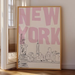 以下が含まれることがあります： クリーム色の背景に大きなライトピンクのブロック体で「NEW YORK」という文字が描かれた、長方形の額入りウォールアートプリント。文字の下には、自由の女神像とニューヨークの街並みが黒い線画で描かれています。