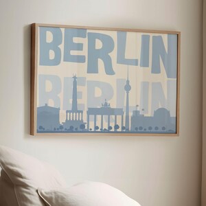 Op de afbeelding: Een ingelijste print met het woord "BERLIJN" in lichtblauw, over een silhouet van de skyline van Berlijn. Het kunstwerk heeft een lichte houten lijst en hangt aan een neutrale muur.