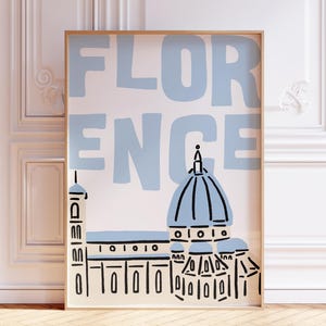 May include: A framed art print featuring the word "FLORENCE" in light blue block letters above a stylized illustration of the Florence Cathedral. The artwork is set against a cream background and framed in a light wood.