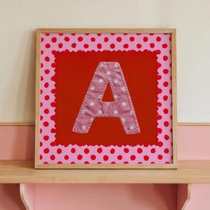 Può includere: Un'opera d'arte incorniciata con la lettera "A" in tessuto a pois rosa, su sfondo rosso con bordo a pois rosa. L'opera è esposta su una mensola in legno chiaro e rosa, creando un'esposizione decorativa e giocosa.